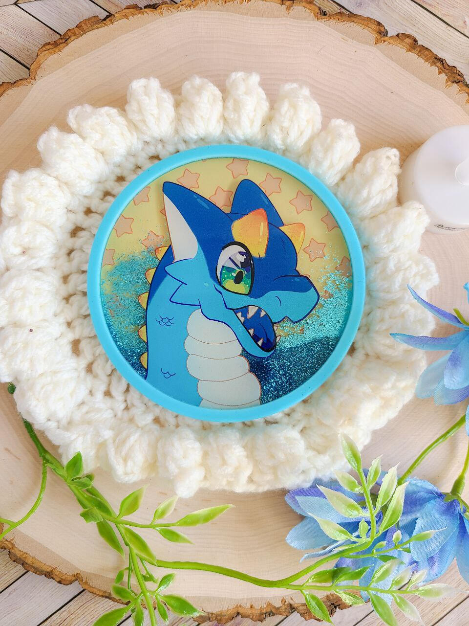 A blue glitter filled coaster sits atop a crocheted place mat with foliage decorating the frame. The coaster depicts a smiling blue dragon with yellow horns.