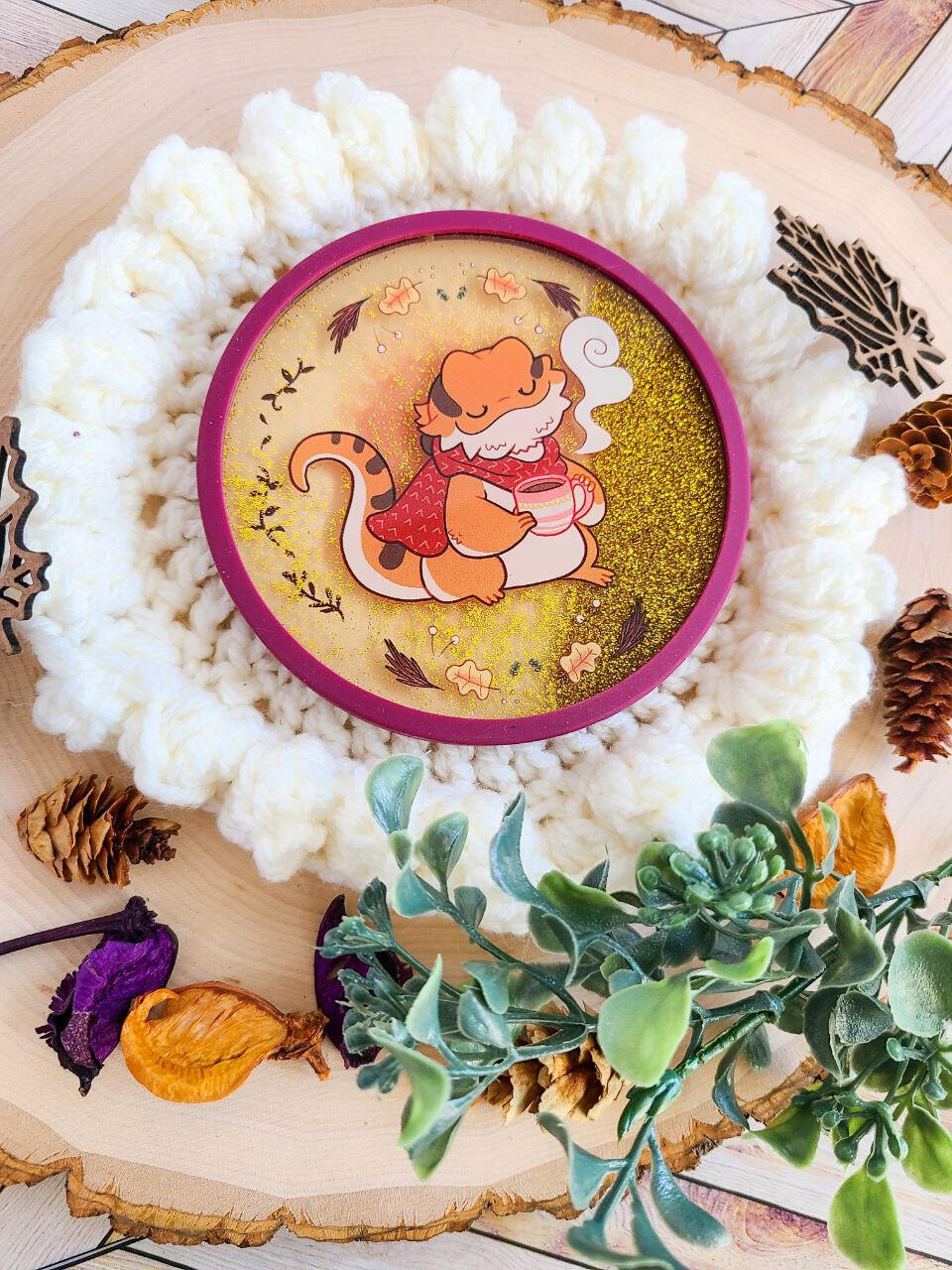 A gold glitter filled maroon coaster sits atop a crocheted place mat with foliage decorating the frame. The coaster depicts a content looking bearded dragon wearing a red scarf and holding a small tea cup.