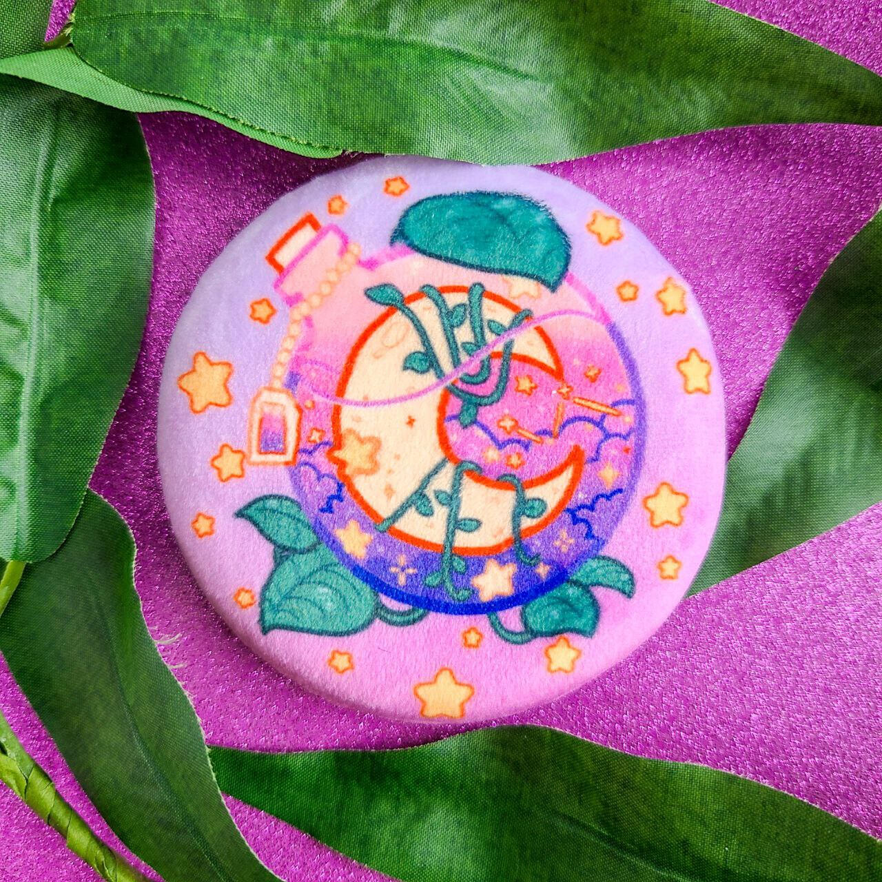 A plush button depicting a round potion bottle containing a crescent moon and clouds. It is wrapped in moonlight trebuii plants on a light pink background.