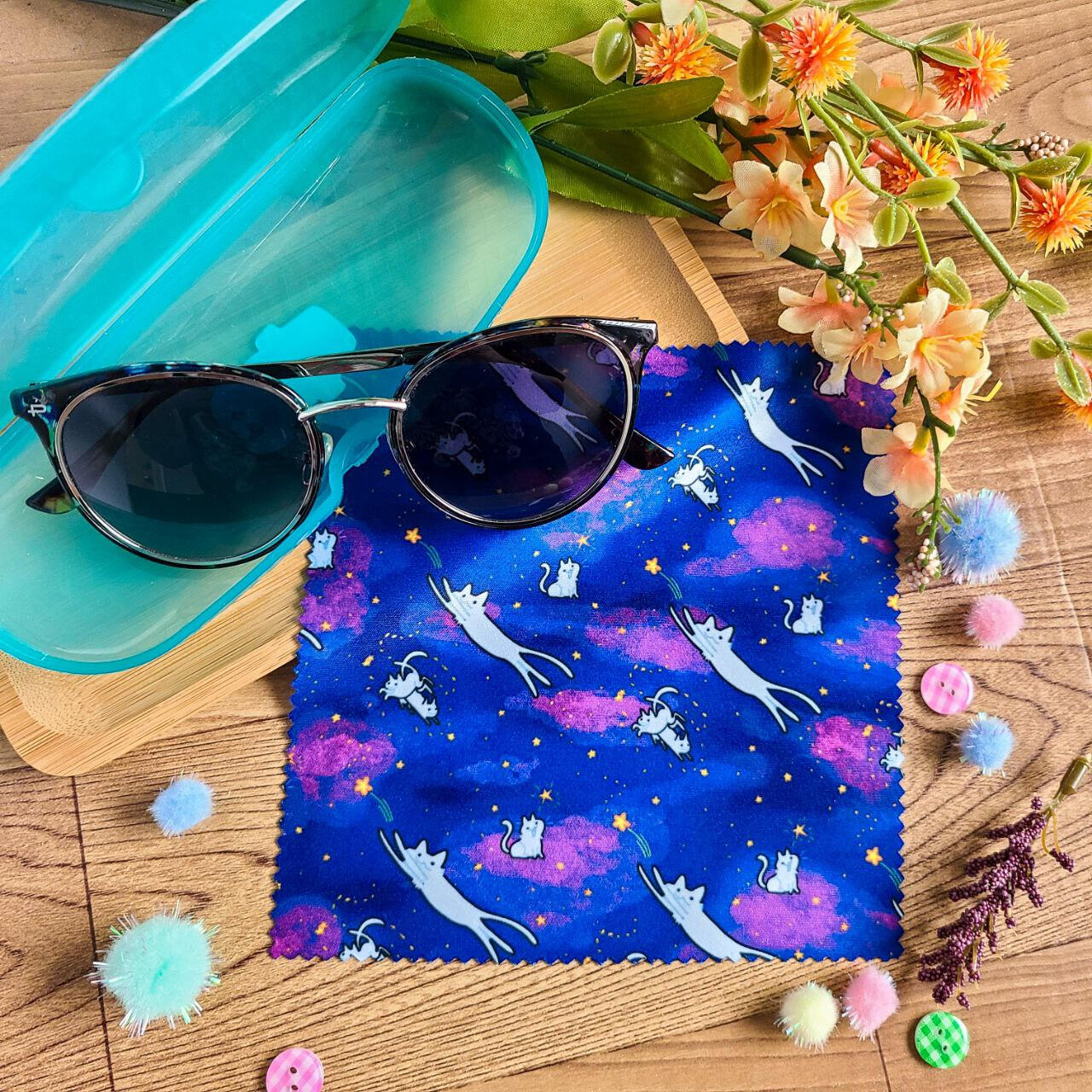 A purple-blue glasses cloth rests on a wooden background with sunglasses placed on top. The cloth depicts a pattern of white cats sprawling across the starry filled sky.
