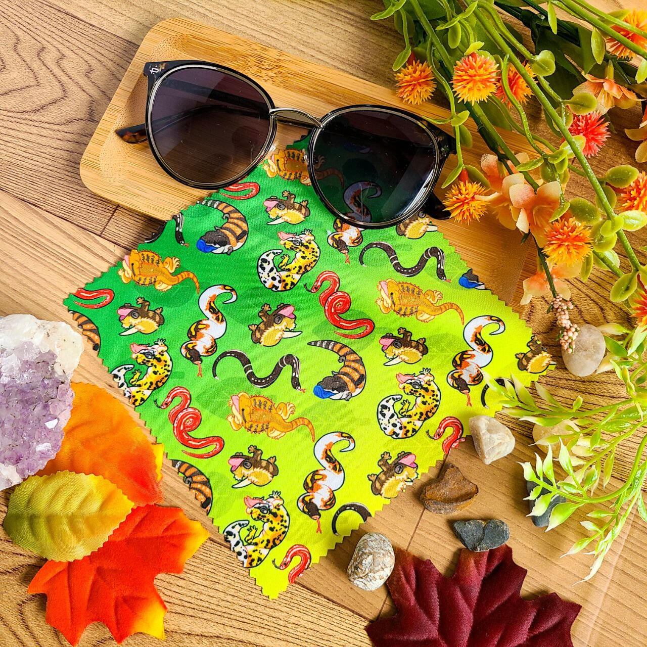 A green and yellow glasses cloth rests on a wooden background with sunglasses placed on top. The cloth depicts a variety of different lizards and snakes. A gecko, skink, bearded dragon, corn snake, king snake, and ball python can be seen.