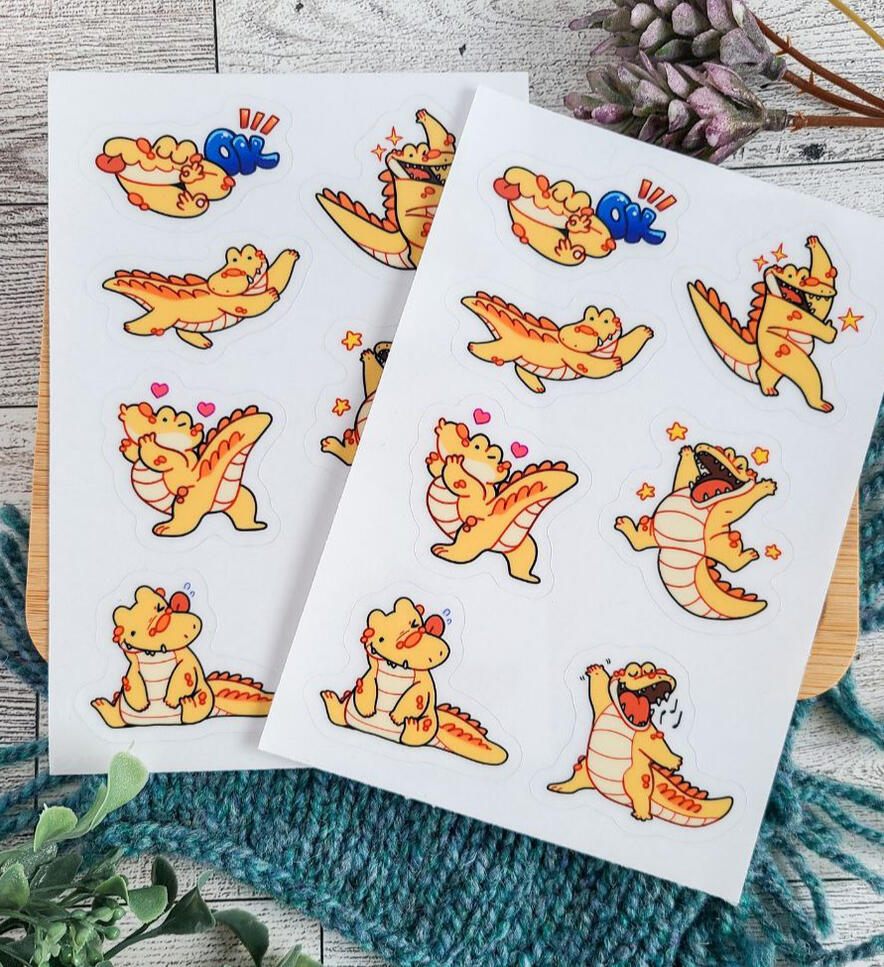 Two sticker sheets depicting my crocodile mascot, Cheddar. She is making eight different poses very energetically.