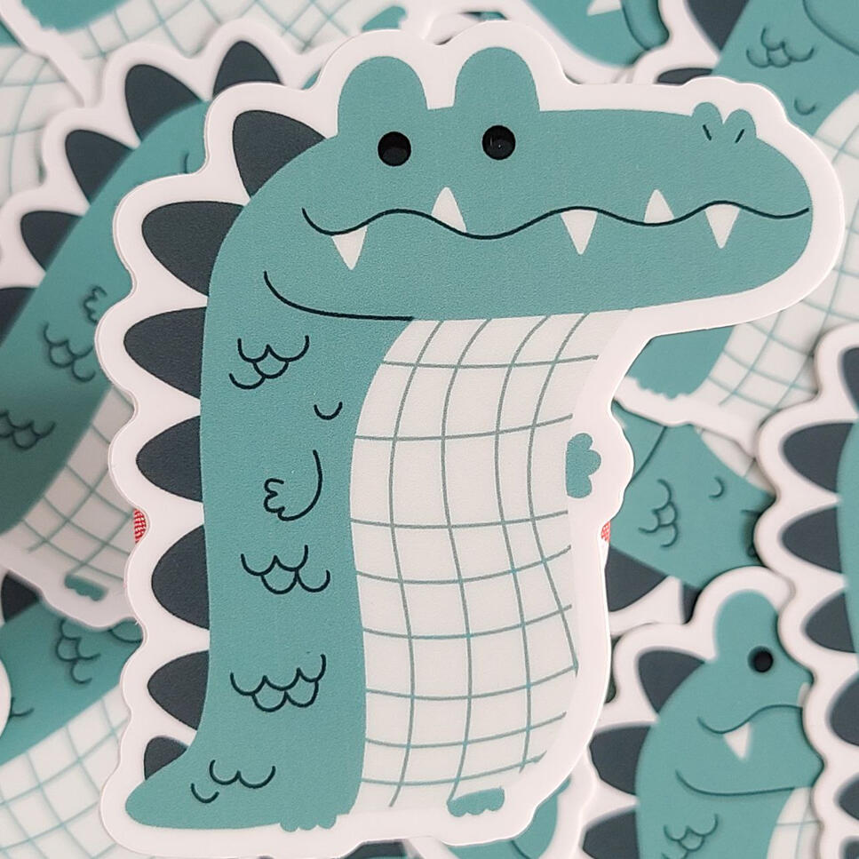 A sticker of a long, green and white alligator / crocodile. It has a wiggly smile with teeth poking out.