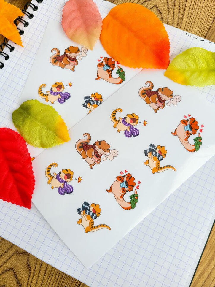 A sticker sheet of eight bearded dragons dressed for fall. Laid on a background of graph paper and surrounded by autumn colored leaves.